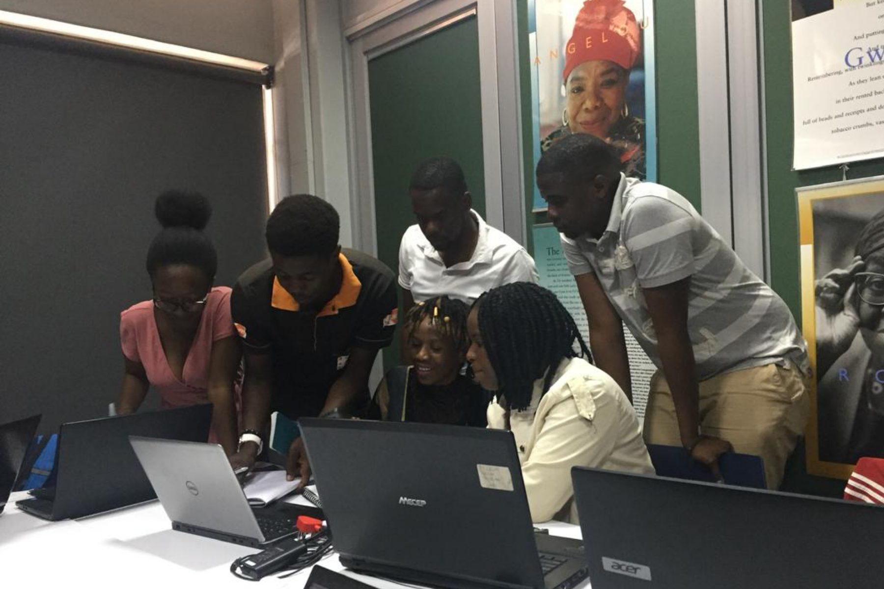 Students gathered around a laptop in a session, one laughing, several leaning in to look at the same screen