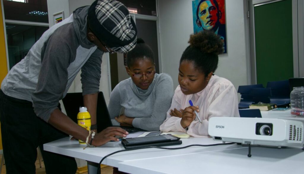 A teaching assistant leaning over a desk explaining something to two students, laptops and notebooks open